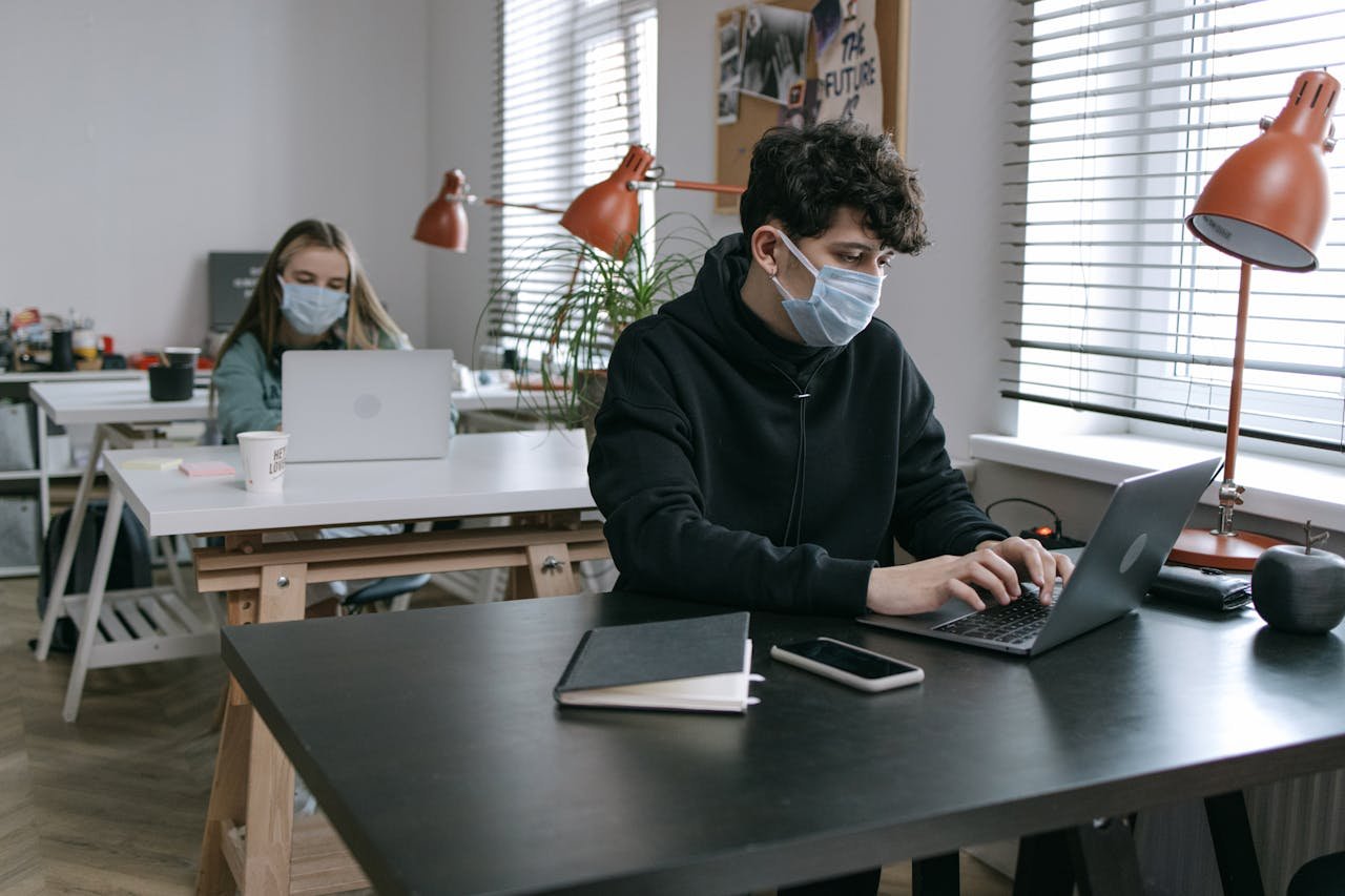 services-04 Two people wearing masks working on laptops in a modern office, maintaining social distance.