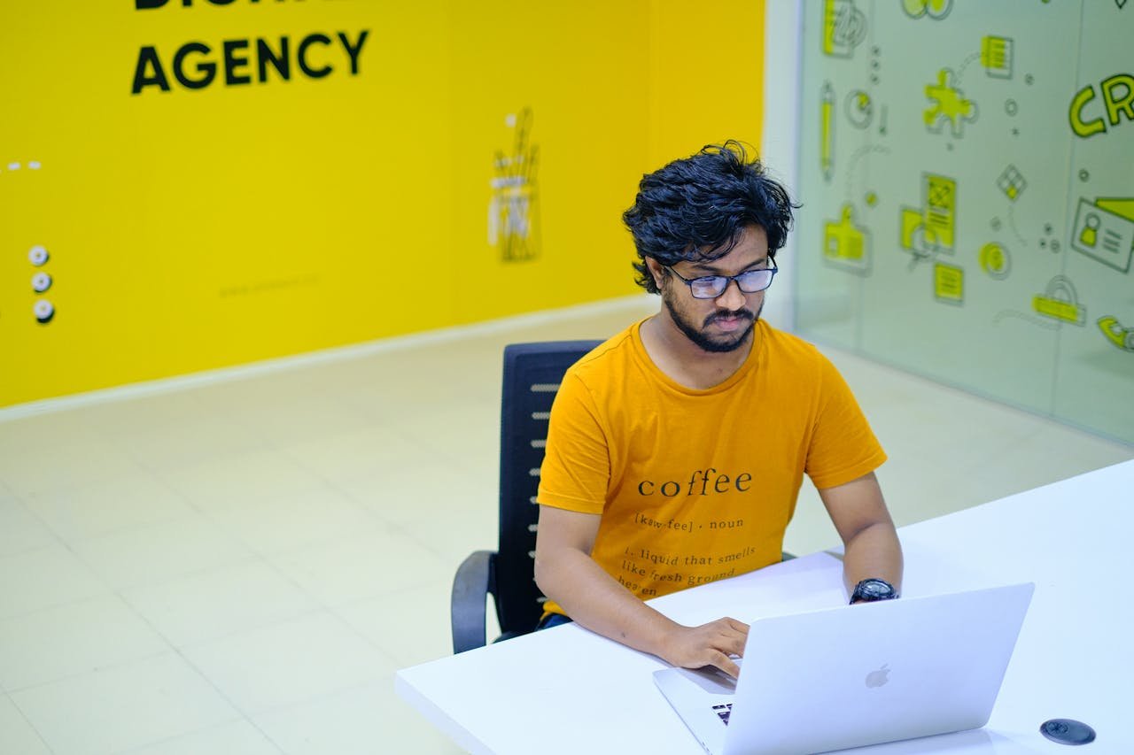 Young man wearing eyeglasses working on laptop in vibrant digital agency office.